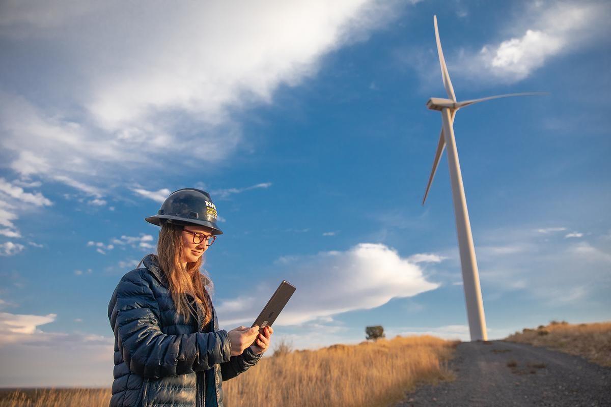 A student wearing a construction hardhat looks at an IPad and stands in front of rotating wind turbine.