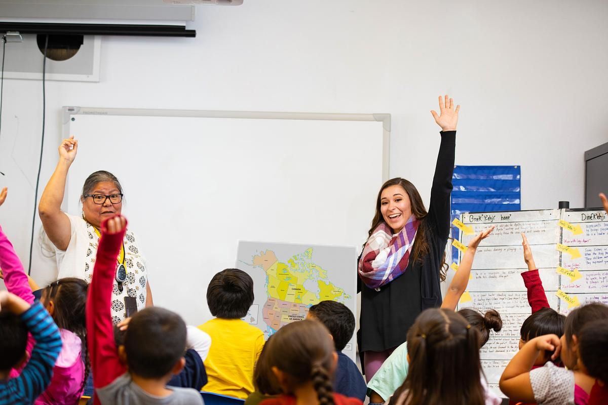 A teacher and student instructor encourages young students to raise their hands in the classroom while standing in front of a whiteboard and map of North America.