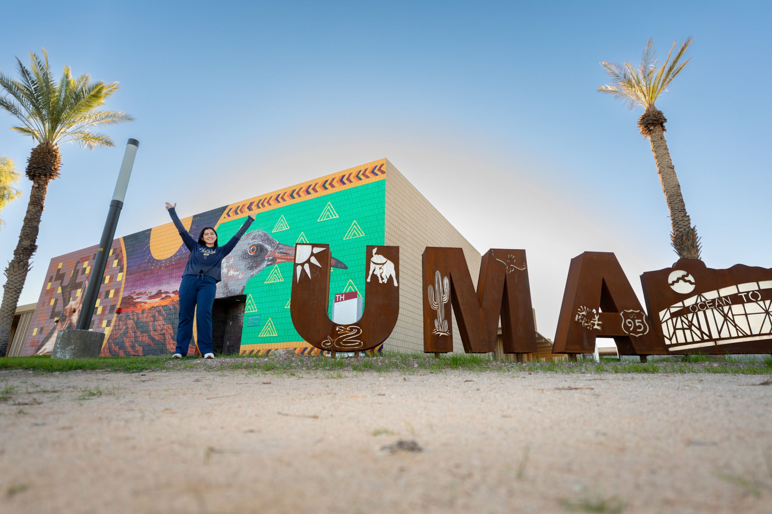 An NAU student makes a Y with her arms in the air next to a sign to spell out Yuma.