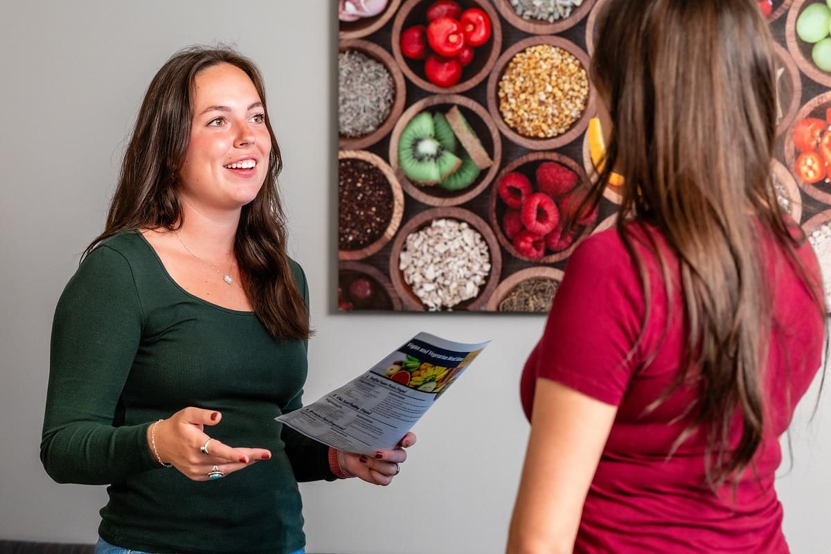 A student in the Nutrition program holds a pamphlet in front of a photograph with assorted fruits and healthy foods.