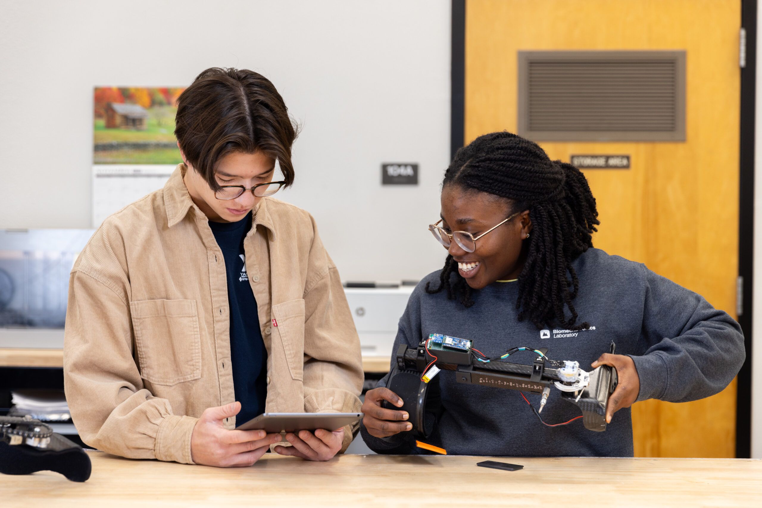 Two students collaborate in a lab, one holding a tablet and the other smiling while working on a robotic device with exposed circuitry.