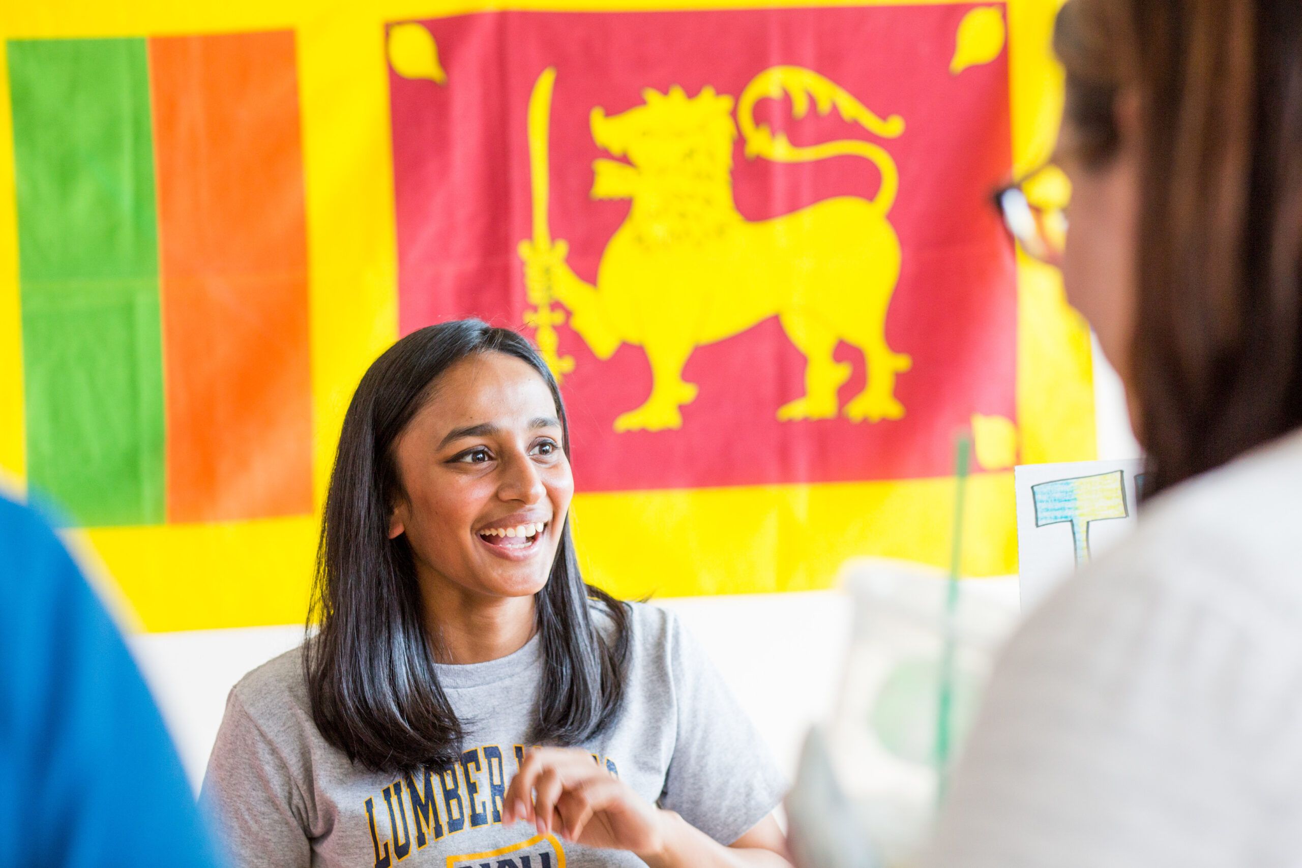 Student with a "Lumberjack" tee shirt is smiling and standing in front of a group of international flags.