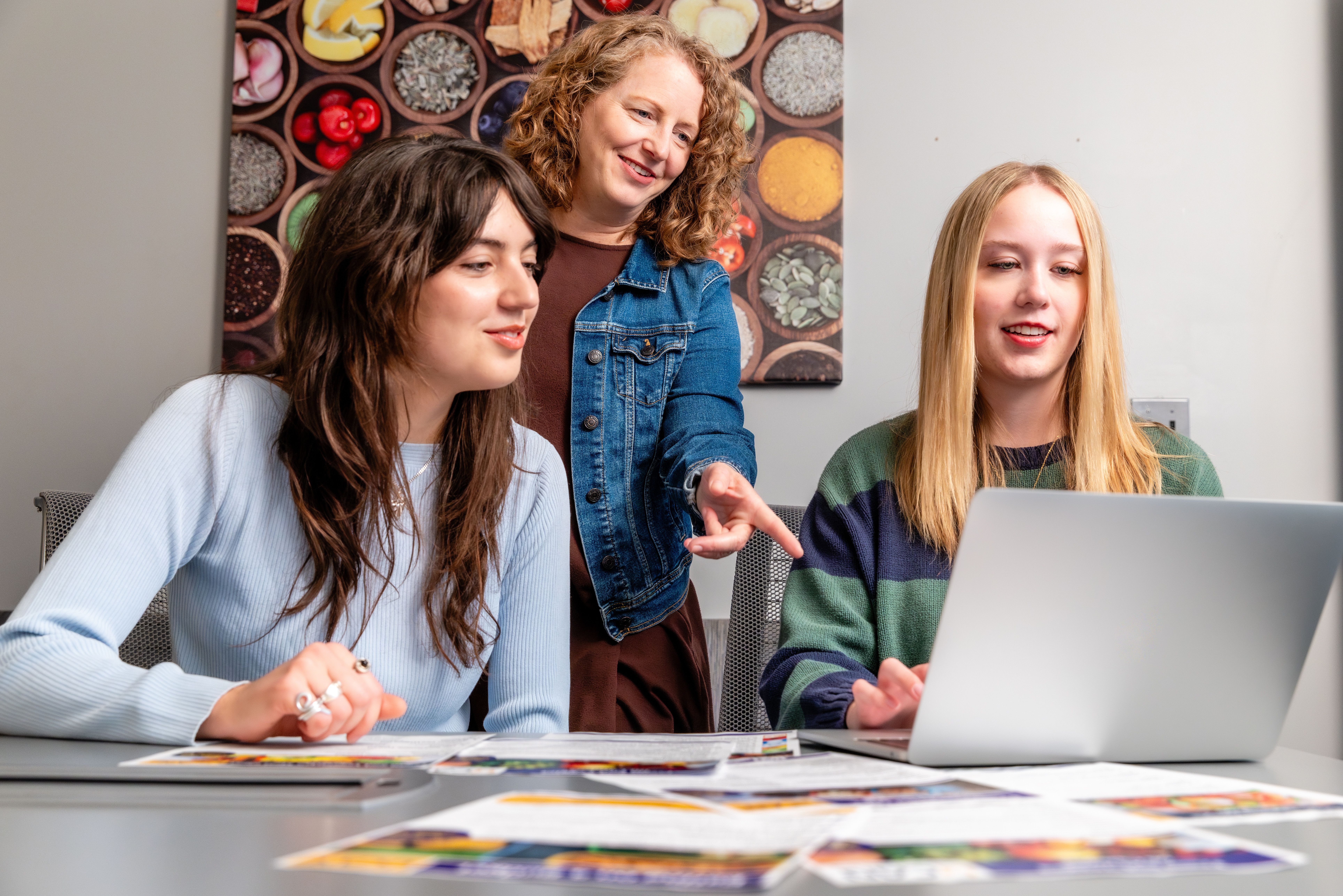 Three individuals sitting at a table, interacting with a laptop computer and some printed materials. The individuals appear to be engaged in discussion or collaborative work.